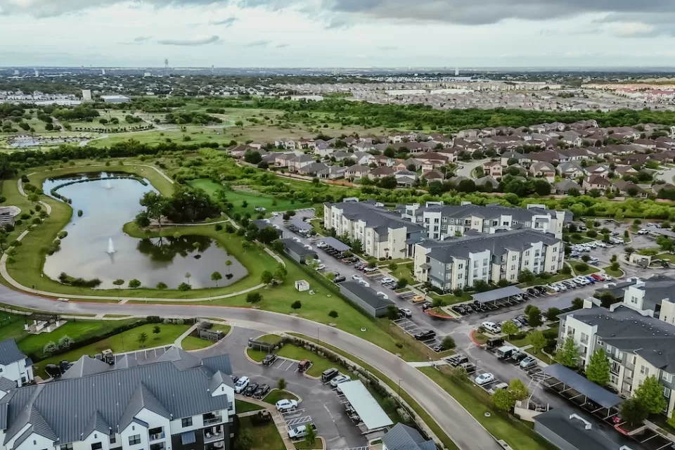 Aerial view of suburban mixed-use development outside Austin, Texas with lake in foreground and complex of apartment buildings.